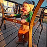 toddler, child, hat, green_sleeves, orange_pants, wooden_deck, metal_table, metal_chair, sunlight, shadow, outdoor, smiling, happy, baby, clothing, handrail, fence, daylight, person, playful