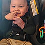 Gordon is registered to the contest to win money with this photo: baby, infant, child, smiling, cheeks, face, hand, fingers, car_seat, seat_belt, buckle, toy, warning_label, portrait, close_up, green_shirt, pants, happy, indoor, cute