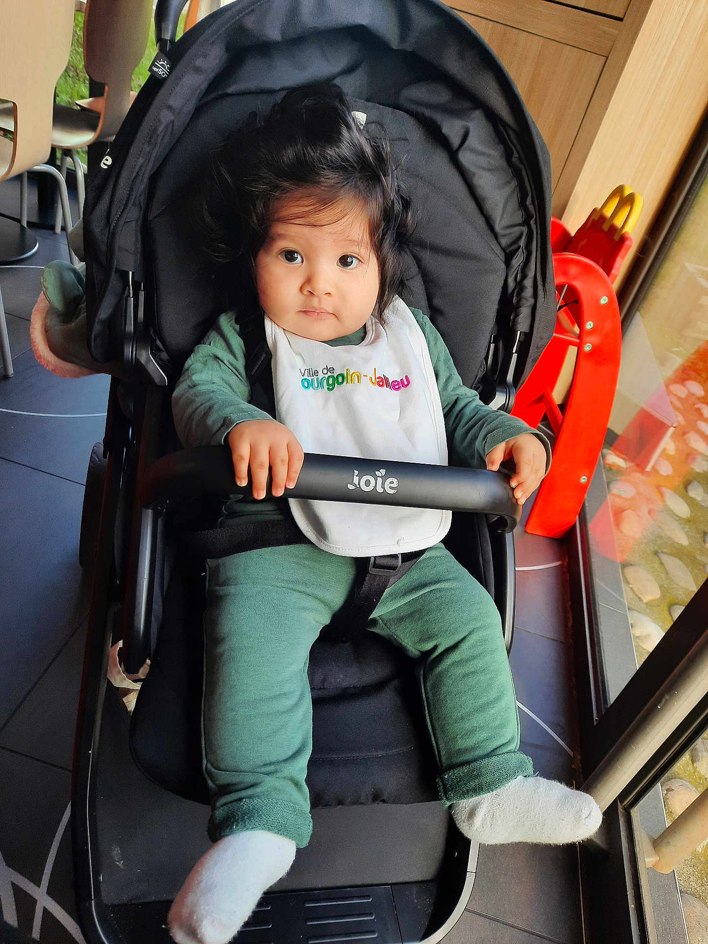 Youness Ben Mena participe au concours pour gagner de l'argent avec cette photo : baby, bib, black_stroller, chair, child, cute, expression, face, floor, green_clothing, hair, hand, indoor, infant, person, safety_bar, sitting, stroller, white_socks, window