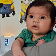 Sebastian is registered to the contest to win money with this photo: baby, infant, child, portrait, minion, cartoon, wall_poster, indoor, person, hand, arm, face, hair, eyes, green_shirt, shoulder, adult_arm, decor, cute, adorable