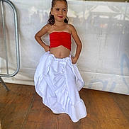 Marie participe au concours pour gagner de l'argent avec cette photo : child, colorful_clothing, confident, cute, event, fashion, girl, hands_on_hips, happy, indoors, long_hair, portrait, posing, red_top, smiling, standing, tent, white_skirt, wooden_floor, young