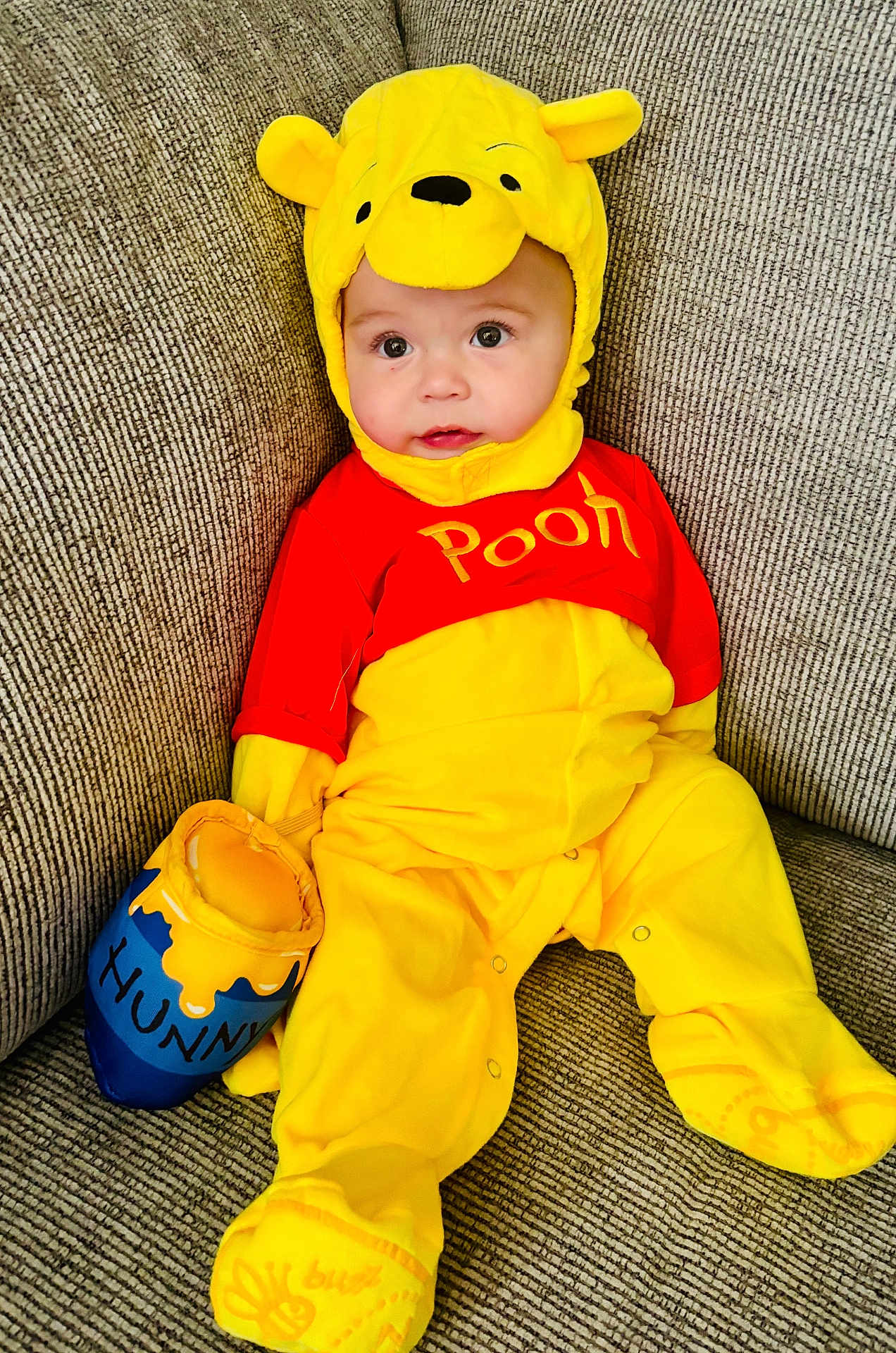 Sutton is registered to the contest to win money with this photo: baby, child, costume, yellow, red, winnie_the_pooh, plush, honey_pot, sofa, couch, seated, cute, indoor, face, clothing, footwear, happy, portrait, infant, soft_toy