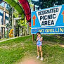 child, girl, swimsuit, water_slide, picnic_area_sign, grass, outdoor, playground, summer, trees, park, smiling, posing, daylight, nature, footwear, sandals, fun, recreation, summer_fun