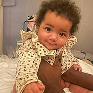 Myrah is registered to the contest to win money with this photo: baby, infant, child, person, smile, curly_hair, ruffled_clothing, hand, bed, blanket, indoor, cozy, cute, patterned_clothes, skin, face, eyes, holding, soft_light, closeup