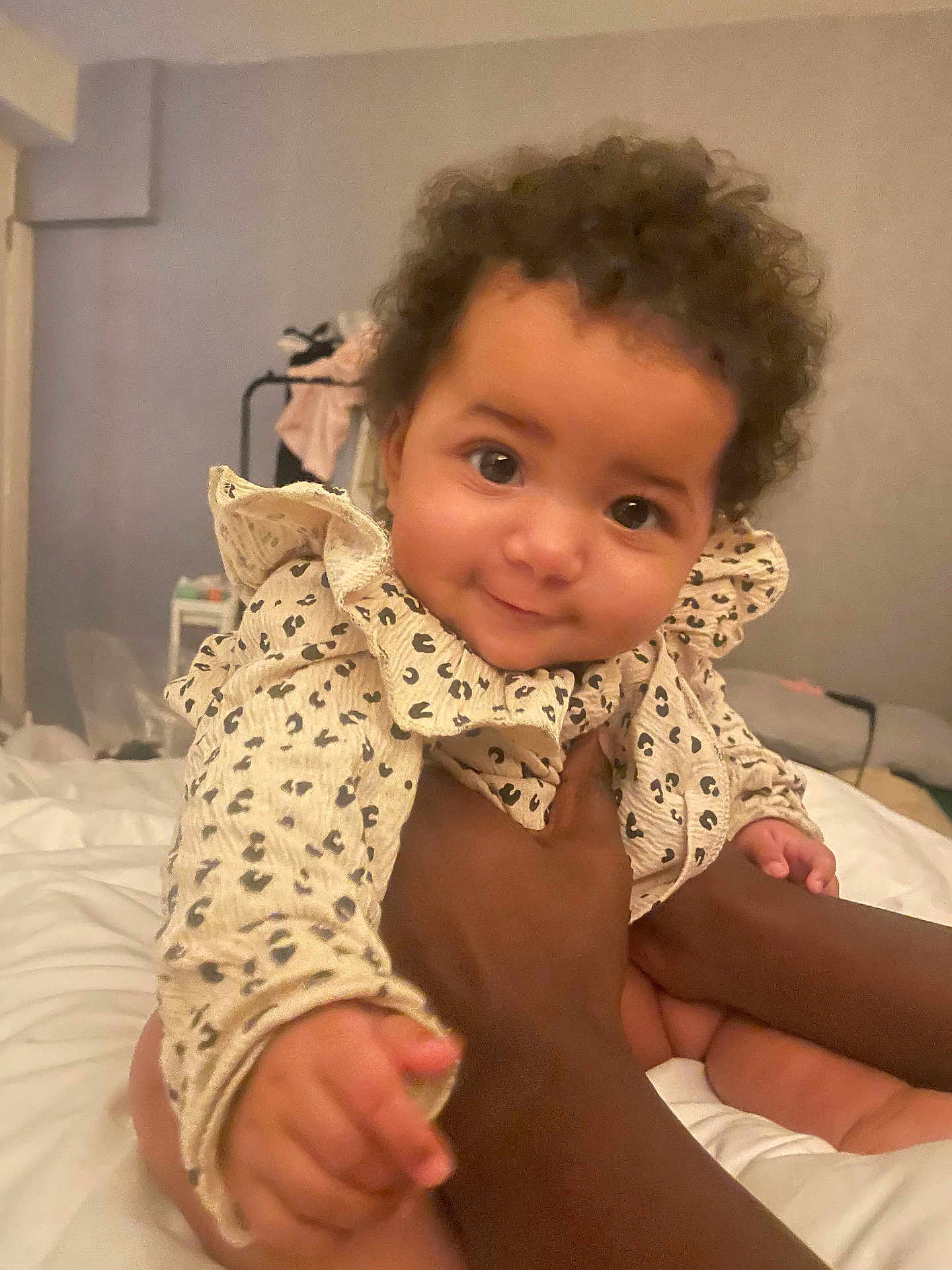 Myrah is registered to the contest to win money with this photo: baby, infant, child, person, smile, curly_hair, ruffled_clothing, hand, bed, blanket, indoor, cozy, cute, patterned_clothes, skin, face, eyes, holding, soft_light, closeup