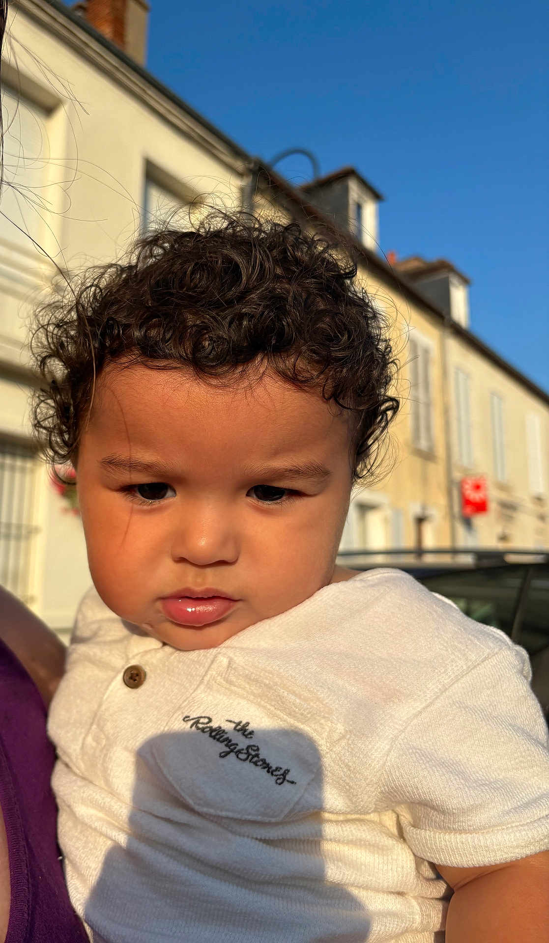 Ismaël a rejoint le concours — aidez-le/la à gagner de superbes lots ! child, toddler, curly_hair, white_shirt, embroidery, outdoor, daylight, portrait, serious_expression, face, person, building, sky, sunlight, shadow, urban, window, clothing, head, close_up