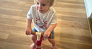 Zachary is registered to the contest to win money with this photo: toddler, child, indoor, balance_bike, wooden_floor, barefoot, shorts, tshirt, blonde_hair, toy, concentration, person, flooring, young_child, riding, handlebars, play, cute, home, casual_clothing
