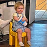 Zachary is registered to the contest to win money with this photo: toddler, child, indoor, yellow_chair, blonde_hair, holding_object, cup, socks, wooden_floor, door, natural_light, wall_art, home_interior, casual_clothing, shorts, short_sleeve_shirt, smiling, seated, person, footwear