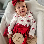 Brielle joined the competition — help win amazing prizes! baby, infant, child, smiling, red_bow, heart_pattern, dress, white_blanket, wooden_sign, five_months, cute, portrait, indoor, happy, young_child, headband, sitting, person, milestone, cozy