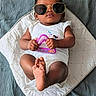 baby, child, sunglasses, toy, white_clothing, cushion, blanket, indoor, portrait, infant, cute, relaxed, person, feet, hand, face, fashion, accessory, playful, laying_down