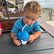 Waylon is registered to the contest to win money with this photo: beach, blue_shirt, casual, child, curly_hair, daylight, drinking, fence, outdoor, people, portrait, restaurant, sand, shark_cup, smiling, straw, table, toddler, umbrella, wood