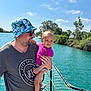 man, child, toddler, boat, water, river, sky, trees, hat, sunglasses, smile, holding, summer, outdoor, nature, casual_clothing, netting, sunlight, happy, family