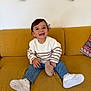 toddler, child, smiling, sitting, couch, yellow_couch, white_sneakers, striped_sweater, blue_jeans, soft_toy, happy, indoor, curly_hair, cute, casual_clothing, playful, portrait, home, furniture, cozy