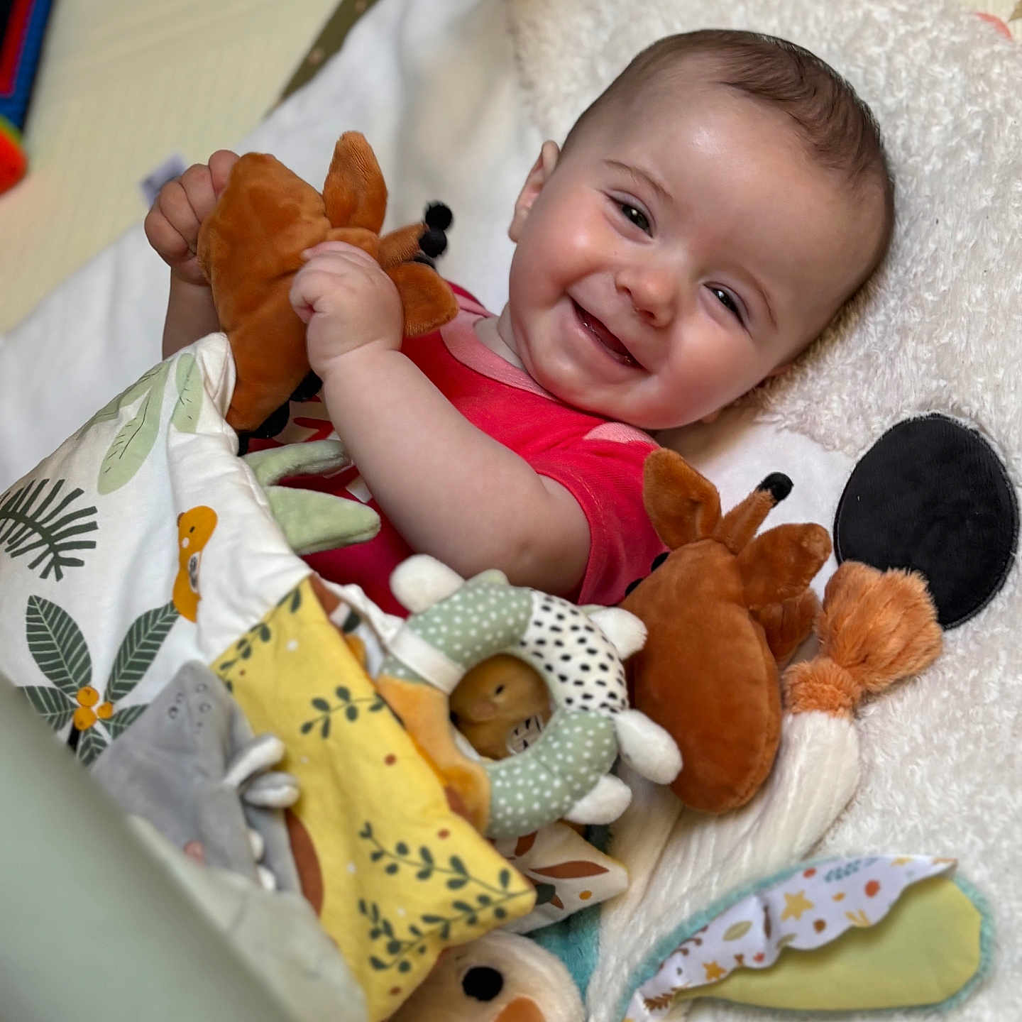 Elliot a rejoint le concours — aidez-le/la à gagner de superbes lots ! baby, smile, plush_toy, blanket, red_clothing, face, child, happy, indoor, soft_texture, cute, infant, playful, colorful, comfort, lying_down, cozy, toy, person, portrait
