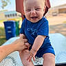baby, background_blur, blue_clothing, casual, child, daylight, face, feet, glass_table, hand_holding, happy, nature, onesie, outdoor, person, recycling_bin, skin, smile, table, tree