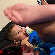 Bryan is registered to the contest to win money with this photo: baby, foot, bottle, blanket, smile, happy, child, indoor, person, skin, blue_blanket, red_blanket, lying_down, playful, cute, infant, hand, face, young, cozy
