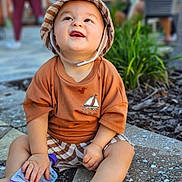 Nayip Andre Inigo is registered to the contest to win money with this photo: toddler, child, baby, hat, bucket_hat, smiling, happy, outdoor, pavement, snack_pouch, clothing, shorts, person, greenery, blurred_background, sitting, cute, sunlight, casual, summer