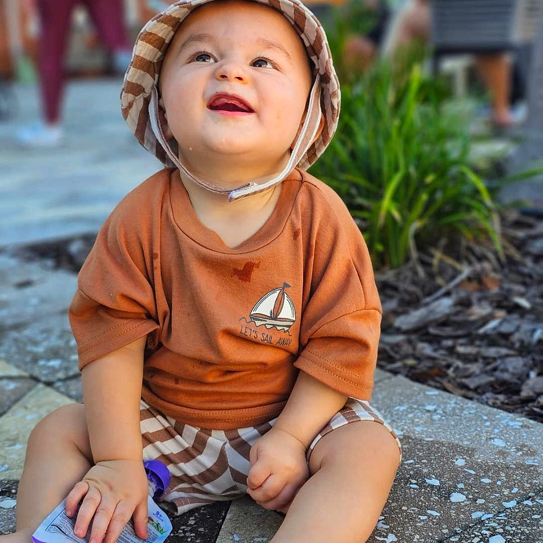 Nayip Andre Inigo is registered to the contest to win money with this photo: baby, blurred_background, bucket_hat, casual, child, clothing, cute, greenery, happy, hat, outdoor, pavement, person, shorts, sitting, smiling, snack_pouch, summer, sunlight, toddler