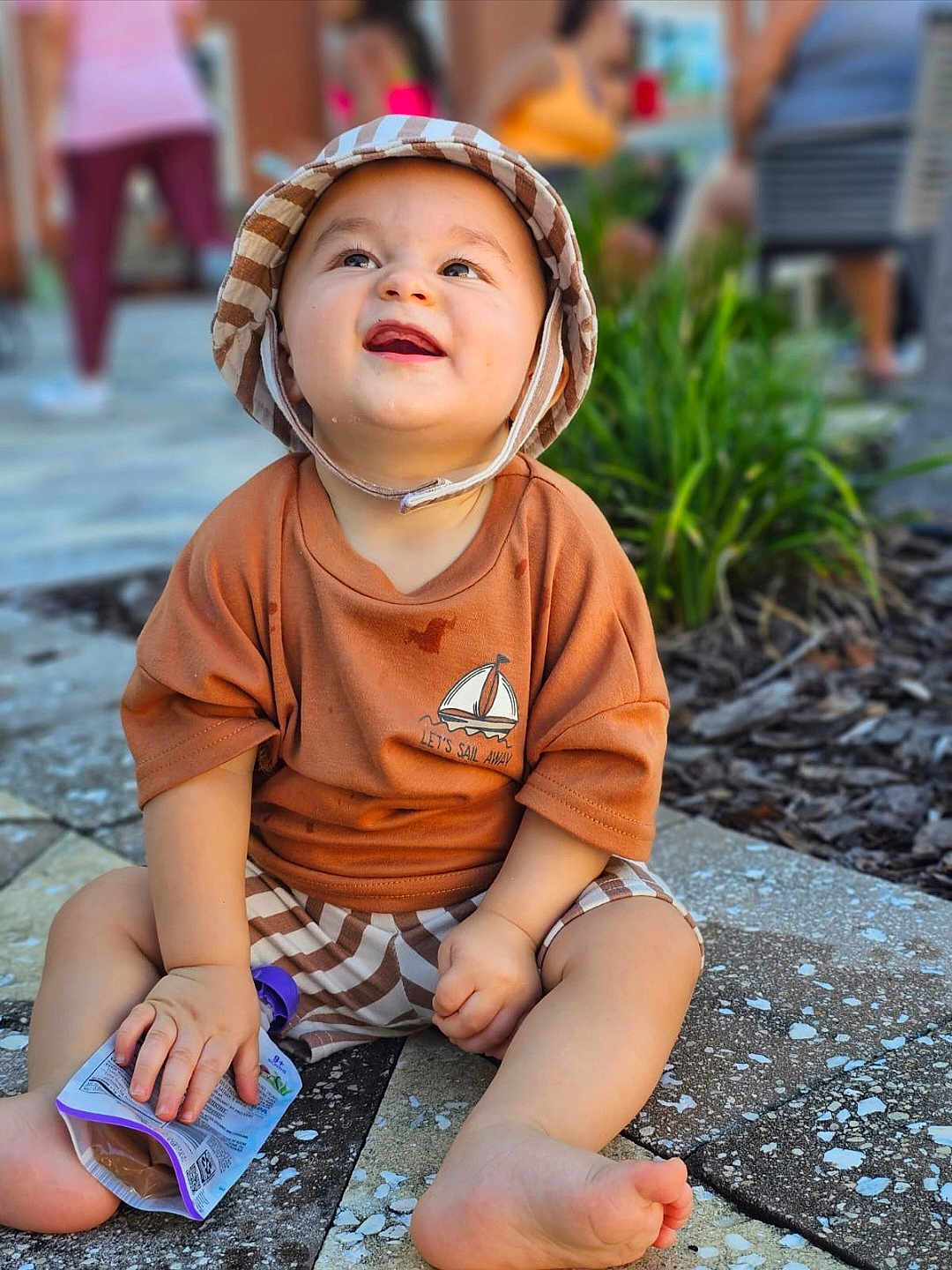 Nayip Andre Inigo is registered to the contest to win money with this photo: toddler, child, baby, hat, bucket_hat, smiling, happy, outdoor, pavement, snack_pouch, clothing, shorts, person, greenery, blurred_background, sitting, cute, sunlight, casual, summer