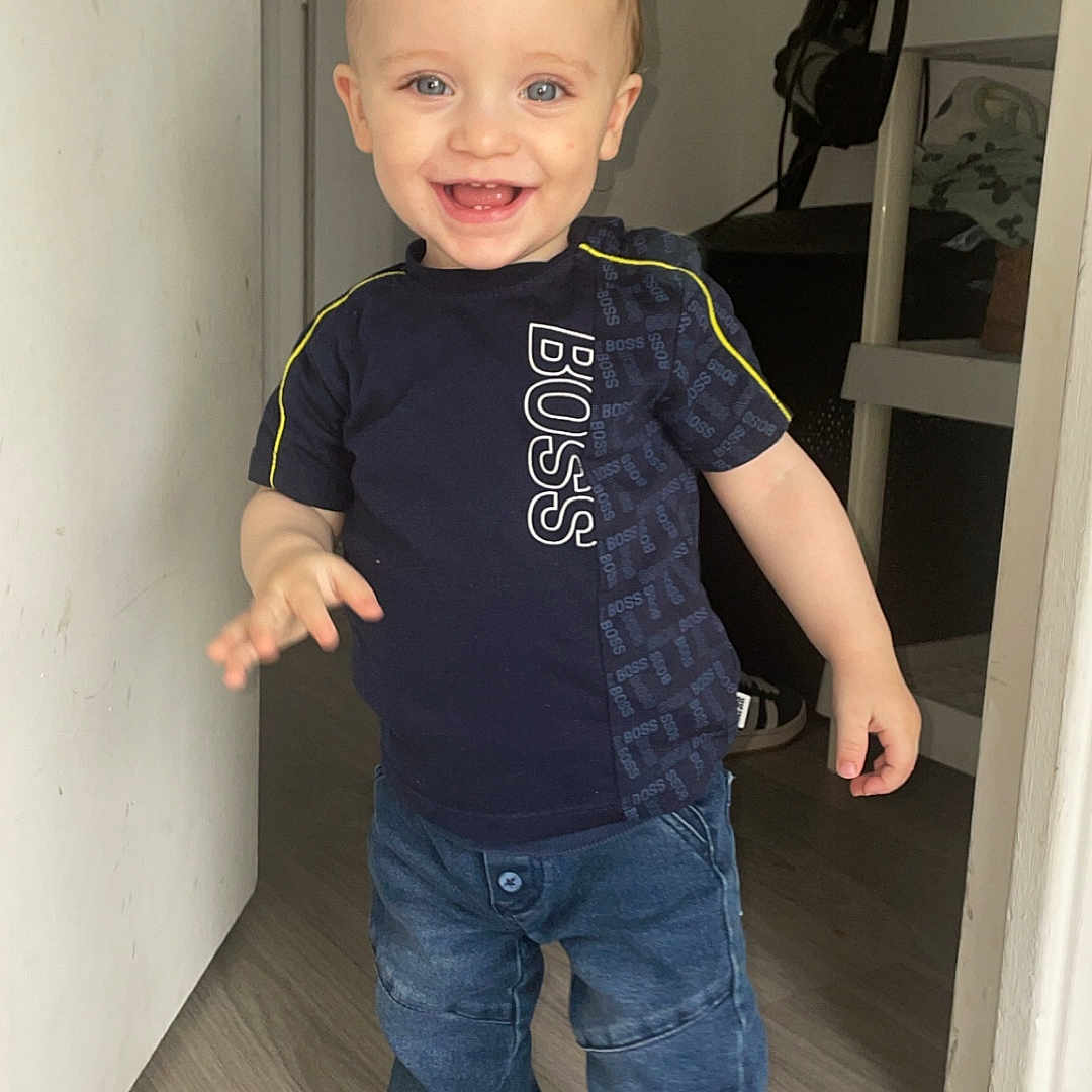 Kaïlan a rejoint le concours — aidez-le/la à gagner de superbes lots ! baby, blue_eyes, casual_clothing, child, denim_pants, doorway, floor, happy, home, indoor, light_skin, person, short_sleeves, smiling, sock, standing, toddler, tshirt, wood_floor, young_child