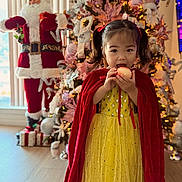 Sophia is registered to the contest to win money with this photo: child, girl, yellow_dress, red_cape, apple, christmas_tree, christmas_decorations, santa_claus_figure, holiday, festive, indoor, smiling, pigtails, lights, ornaments, wooden_floor, celebration, portrait, cute, holiday_spirit