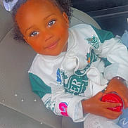 Liyah is registered to the contest to win money with this photo: child, toddler, car_seat, jacket, white, green, bow, pigtails, curly_hair, smile, holding, red_container, sparkles, indoors, cute, portrait, clothing, happy, person, seat