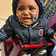 Antonio is registered to the contest to win money with this photo: toddler, child, smiling, black_jacket, hooded_jacket, stickers, face_stickers, high_chair, wooden_chair, camo_pants, indoor, happy, cute, baby, person, seated, clothing, portrait, cheerful, young_child