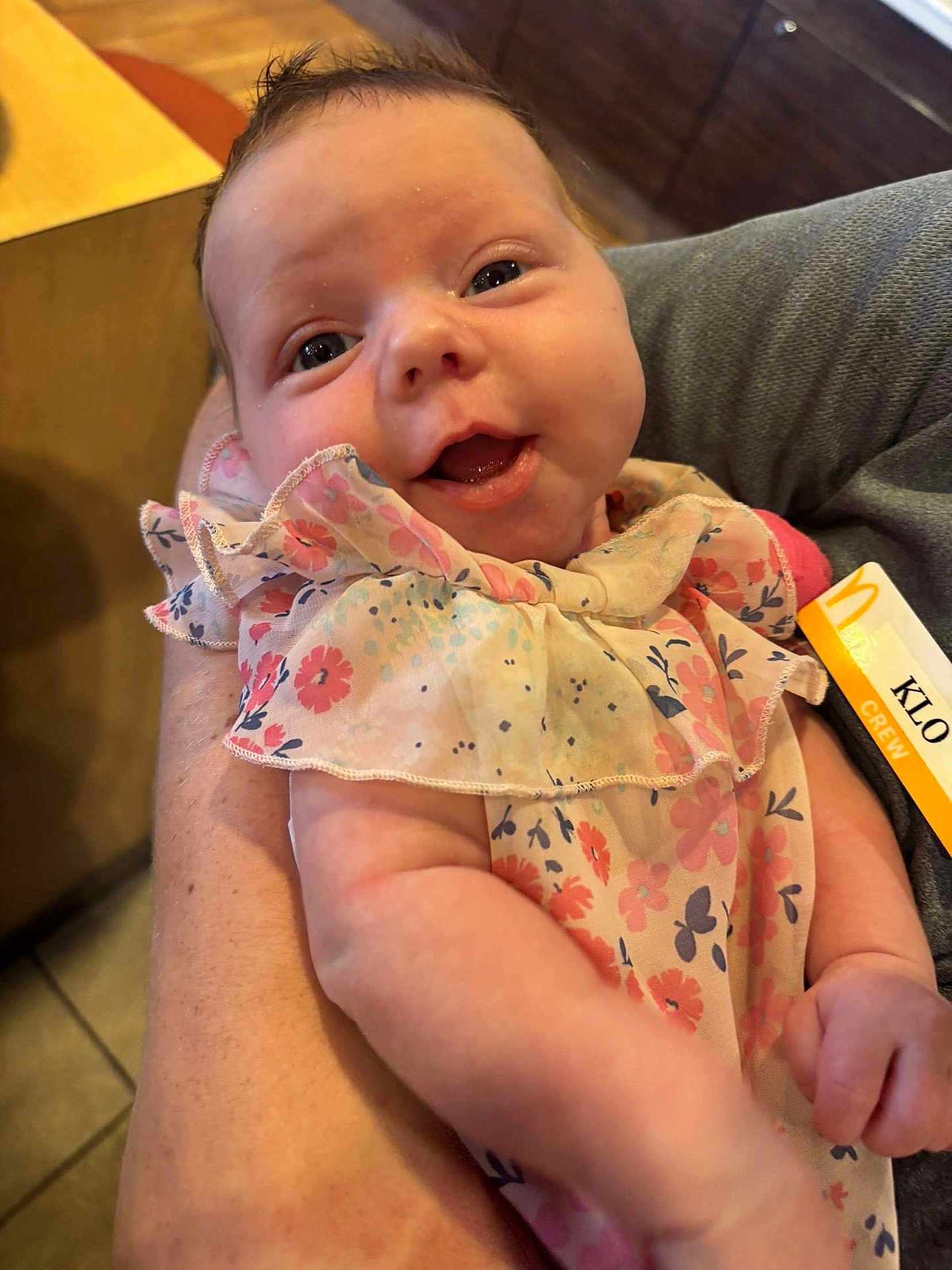 Arizona is registered to the contest to win money with this photo: baby, infant, smiling, face, arm, clothing, floral_dress, person, indoor, holding, human_skin, cute, happy, closeup, child, newborn, portrait, warm_light, fabric, toddler