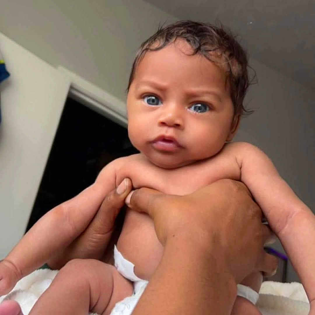 Eric is registered to the contest to win money with this photo: baby, infant, child, portrait, face, eyes, wide_eyed, serious_expression, curly_hair, diaper, being_held, hands, arms, blanket, indoor, home, doorway, close_up, photography, adorable