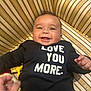 baby, smiling, infant, black_clothing, text_on_clothing, striped_cushion, happy, face, hand, person, cute, child, indoors, portrait, young_child, joyful, baby_girl, baby_boy, sitting, playful