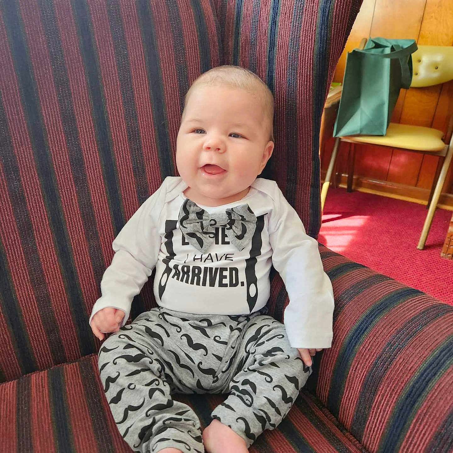 Harlan joined the competition — help win amazing prizes! baby, child, smiling, sitting, armchair, striped_fabric, clothing, onesie, barefoot, indoor, red_carpet, wood_paneling, furniture, person, cute, happy, infant, home, portrait, playful