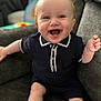 baby, child, smiling, blue_eyes, toothy_grin, navy_outfit, short_sleeves, couch, indoor, happy, cute, infant, person, sitting, comfortable, light_skin, curly_hair, casual_clothing, portrait, joyful