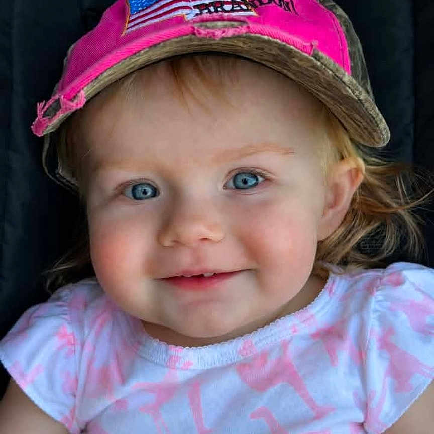 Rebekah joined the competition — help win amazing prizes! toddler, child, baby, portrait, smiling, face, blue_eyes, pink_hat, baseball_cap, pink_shirt, dinosaur_pattern, blonde_hair, rosy_cheeks, sitting, close_up, indoor, dark_background, photograph, happy, cute