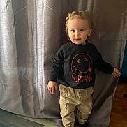 Lyzio participe au concours pour gagner de l'argent avec cette photo : beige_pants, black_sweatshirt, cargo_pants, casual_clothing, child, curly_hair, curtain, expression, flooring, home, indoor, light, looking_at_camera, nirvana_logo, person, portrait, sock, standing, toddler, young_child