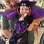 Cain is registered to the contest to win money with this photo: baby, child, man, costume, bat_costume, purple, black, smiling, indoor, person, holding, facial_hair, beard, mustache, plant, furniture, chair, home, cute, happy
