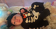 Everleigh is registered to the contest to win money with this photo: baby, infant, child, portrait, lying_down, skeleton_costume, black_clothing, hat, smiling, wide_eyed, face, hands, plush_toy, stuffed_animal, blanket, crib, nursery, indoor, cozy, photography