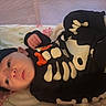 baby, black_clothing, blanket, child, cozy, crib, face, hands, hat, indoor, infant, lying_down, nursery, photography, plush_toy, portrait, skeleton_costume, smiling, stuffed_animal, wide_eyed
