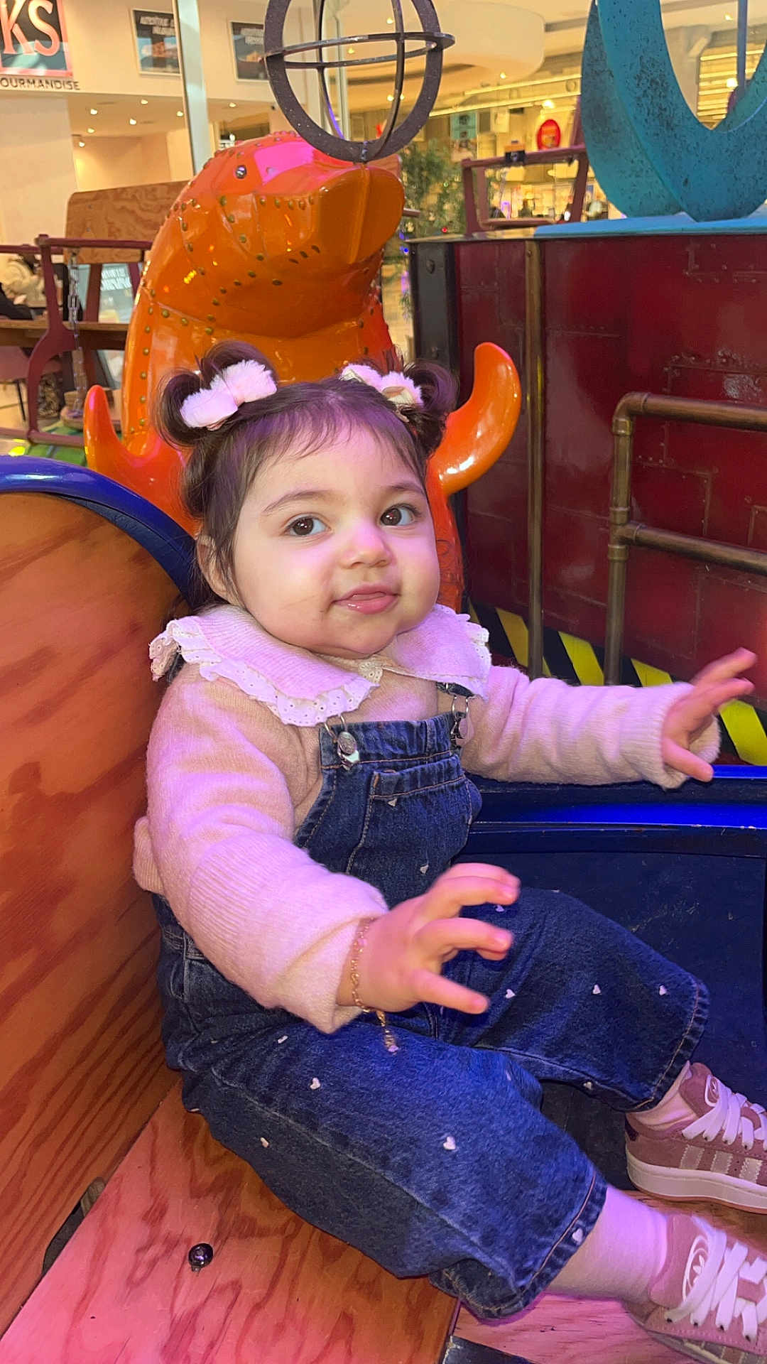 Kiana a rejoint le concours — aidez-le/la à gagner de superbes lots ! child, toddler, baby, face, smile, denim_overalls, pink_sweater, hair_buns, bow_hair, bracelet, sneakers, wooden_seat, amusement_ride, mall_interior, colorful, portrait, sitting, cute, playful, indoor