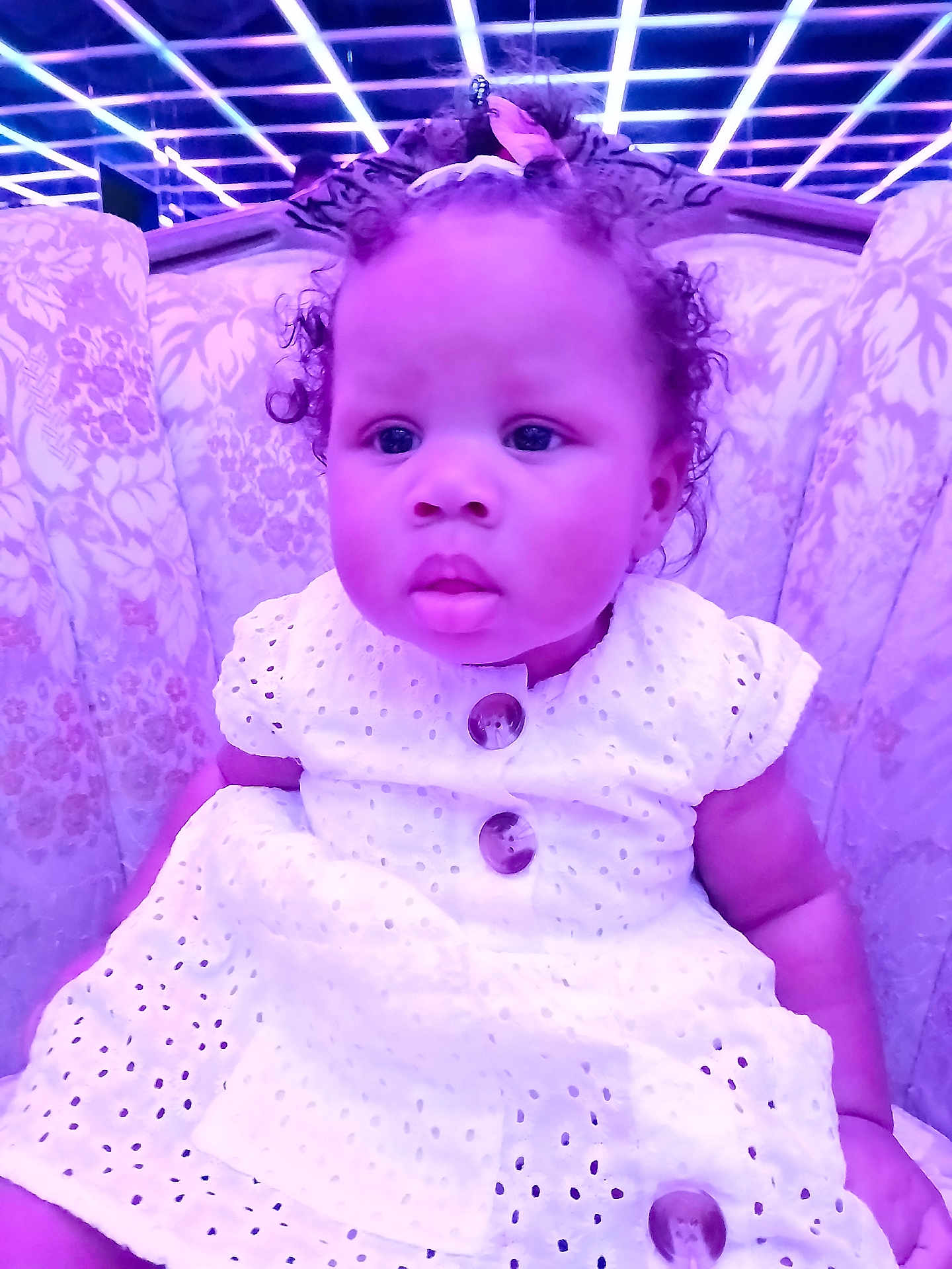 Mckayla is registered to the contest to win money with this photo: baby, child, curly_hair, white_dress, bow, couch, floral_pattern, indoor, portrait, seated, face, eyes, cute, infant, pink_bow, lighting, purple_tint, furniture, expression, young_child