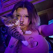 Stormii is registered to the contest to win money with this photo: person, cat, selfie, indoor, purple_light, tabby_cat, piercing, septum_ring, flower_necklace, tank_top, cleavage, tattoo, long_hair, pet, paw, whiskers, tv_screen, bedroom, portrait, jewelry
