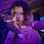 person, cat, selfie, indoor, purple_light, tabby_cat, piercing, septum_ring, flower_necklace, tank_top, cleavage, tattoo, long_hair, pet, paw, whiskers, tv_screen, bedroom, portrait, jewelry