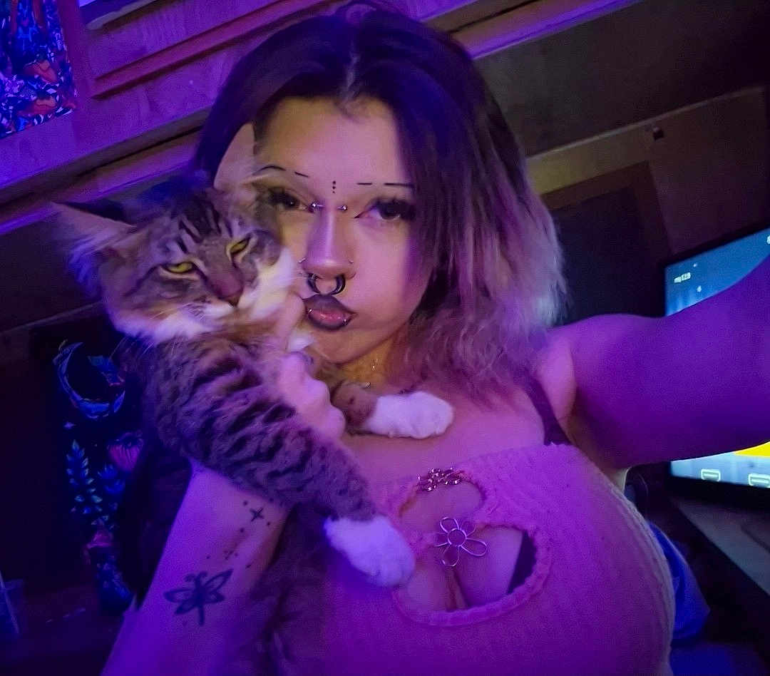 Stormii is registered to the contest to win money with this photo: person, cat, selfie, indoor, purple_light, tabby_cat, piercing, septum_ring, flower_necklace, tank_top, cleavage, tattoo, long_hair, pet, paw, whiskers, tv_screen, bedroom, portrait, jewelry