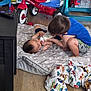 child, baby, feeding, bottle, indoor, floor, mattress, toy, tricycle, blanket, blue_shirt, shorts, wood_floor, playtime, sibling, bonding, casual, cute, home, toddler