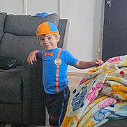 Julian joined the competition — help win amazing prizes! child, boy, smiling, blue_clothing, orange_hat, indoor, couch, blanket, colorful, wood_floor, living_room, person, casual_pose, shorts, furniture, buttoned_shirt, badge, leaning, hat, happy