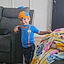 child, boy, smiling, blue_clothing, orange_hat, indoor, couch, blanket, colorful, wood_floor, living_room, person, casual_pose, shorts, furniture, buttoned_shirt, badge, leaning, hat, happy