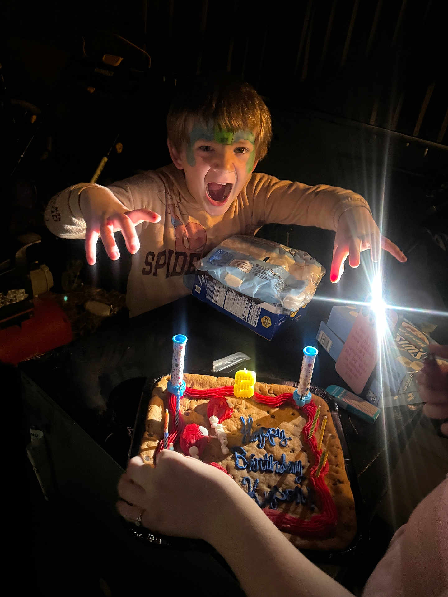 Kyson is registered to the contest to win money with this photo: child, face_paint, birthday, cookie_cake, candles, light, dark, hands, celebration, spider_man_shirt, table, party, excitement, expression, indoor, person, face, food, fun, smile
