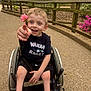 Sawyer is registered to the contest to win money with this photo: child, boy, wheelchair, flower, pointing, black_shirt, outdoor, park, pathway, fence, greenery, happy, smiling, shorts, socks, daylight, people, nature, trees, playful