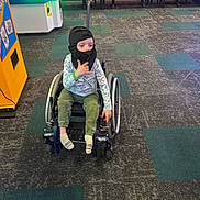 Sawyer is registered to the contest to win money with this photo: child, wheelchair, balaclava, arcade, carpet, hand_gesture, casual_clothing, indoor, flooring, bench, table, game_machine, person, socks, green_bracelet, camouflage_pants, long_sleeve_shirt, play_area, young, seated