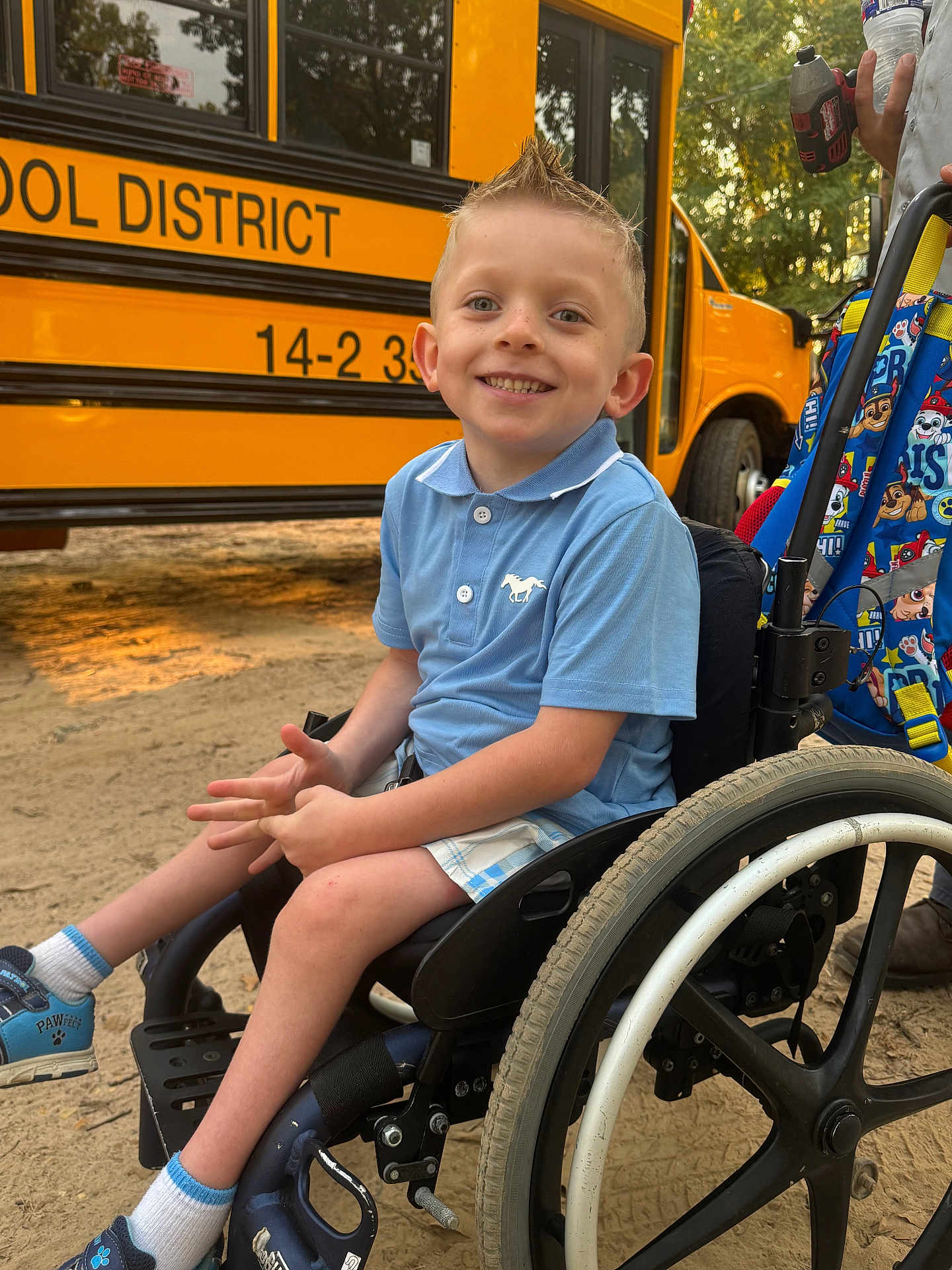 Sawyer joined the competition — help win amazing prizes! child, boy, wheelchair, school_bus, blue_shirt, shorts, smiling, outdoor, backpack, pavement, daylight, person, footwear, socks, vehicle, happy, hair_style, school, hand, wheel