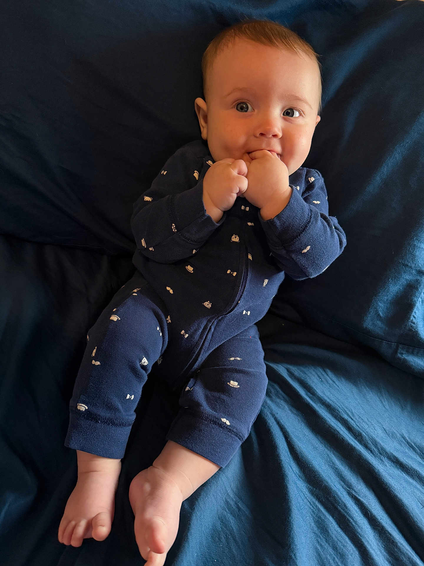 Mason is registered to the contest to win money with this photo: baby, infant, child, onesie, blue_clothing, barefoot, bed, bedroom, fingers_in_mouth, happy, cute, lying_down, skin, head, face, toes, smile, person, indoors, soft_lighting